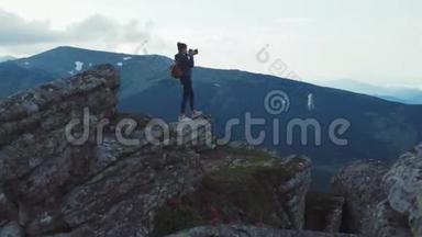 年轻时尚的女摄影师带着背包站在高高的岩石窗台上，拿着相机，俯瞰绿色的景色
